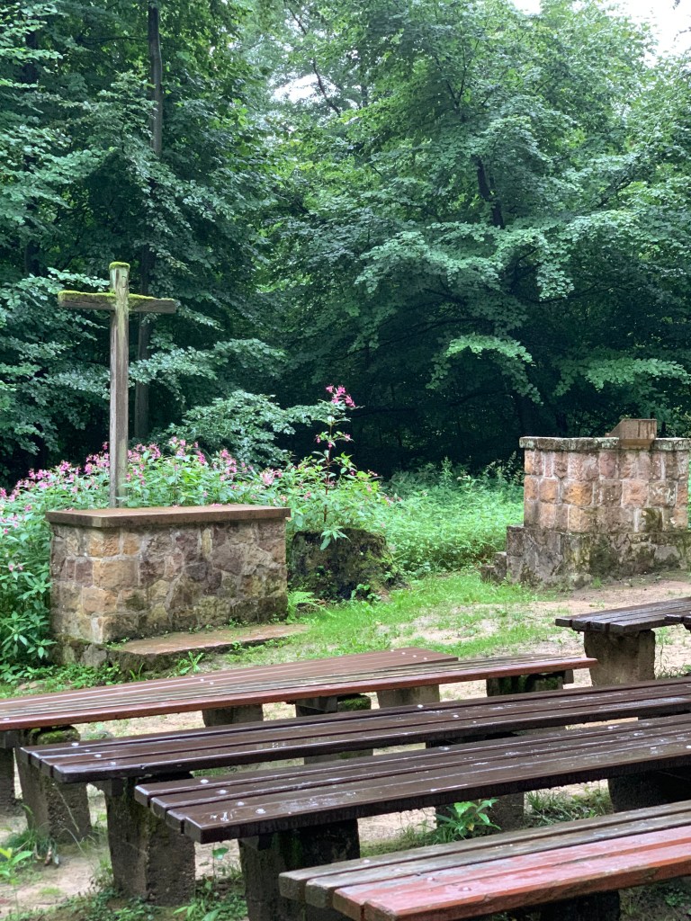 Waldkirche (Kirche im Wald Open Air): Vorne ein Altar mit Holzkreuz, daneben eine Kanzel. Sie und der Altar sind aus Bruchsteinen. Davor Bänke ohne Lehne aus Holz mit in den Boden eingelassenen Betonfüßen. Das Ganze auf leichter Schräge, im Hintergrund Laubwald und überall Regen. 