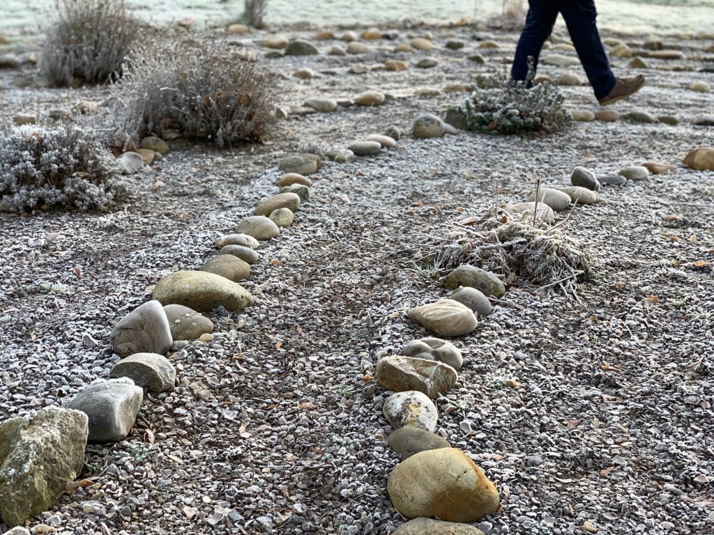 Ausschnitt eines Labyrinths aus Steinen gelegt, dazwischen Kies und mit Raureif überzogen. Am oberen Bildrand sieht man die Beine eines Mannes ausschreiten.