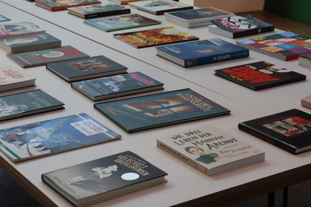 Über einen Teil der Tischplatte mit den Comics drauf fotografiert. Man sieht an die 22 Stück, also etwa die Hälfte. In der Mitte des Tisches sieht man längs an der Ritze, dass der Tisch aus mehreren Elementen zusammengeschoben ist.