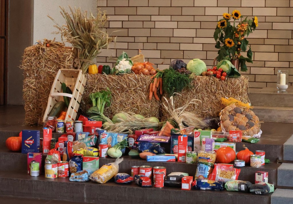 Erntegaben (Obst, Gemüse, Konserven) im Altarraum auf, vor und um drei Strohballen herumdrapiert.