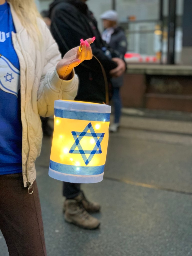 Eine selbst gebastelte Laterne mit der Flagge Israels als Motiv. Gehalten von einer Frauenhand mit langen pinken Fingernägeln, der Ringfinger aber in golden. Sie trägt außerdem ein Israel-T-Sgirt unter der Jacke und eine braune Hose. Ich habe den Kopf bewusst nicht fotografiert. Im Hintergrund verschwommen zwei weitere Teilnehmer und die Kante der Straßenbahnhaltestelle gegenüber der Synagoge.