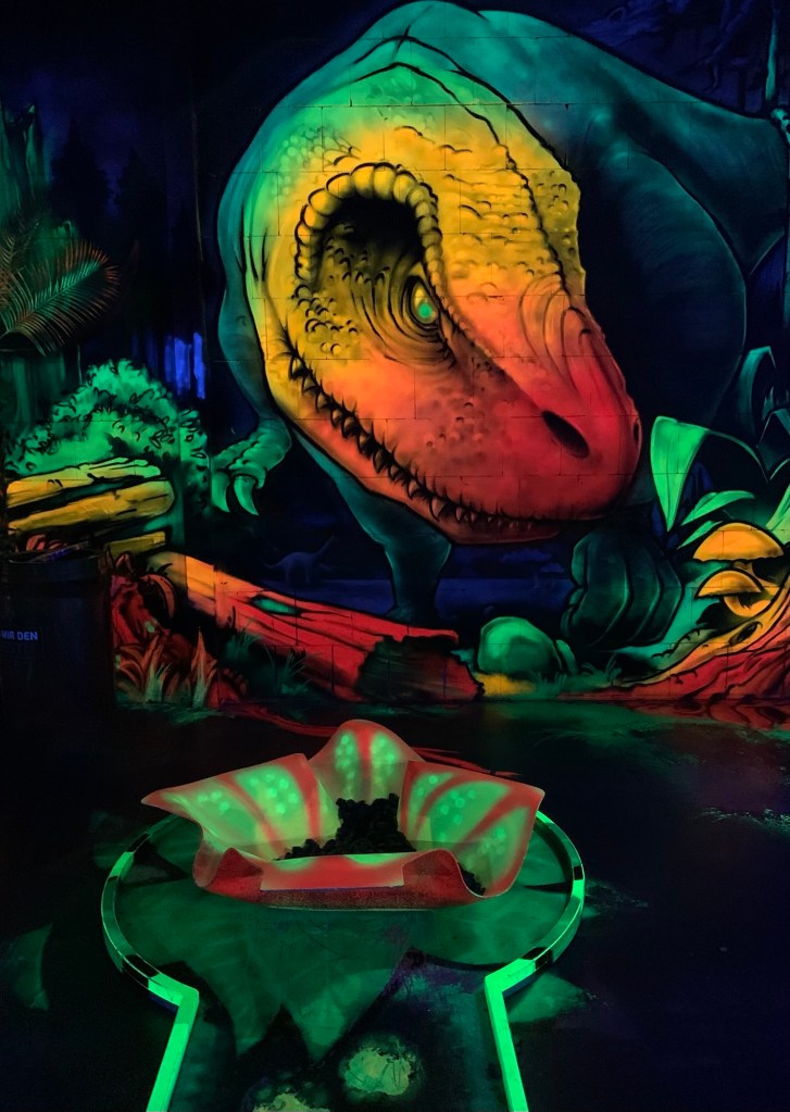 Ein nach vorne gebeugter Tyrannosaurus Rex an der Wand. Davor ein Kelch aus Blättern mit braunem Zeugs drin. Der Kelch steht an der Spitze einer Minigolfbahn, die aussieht, wie ein Schlüsselloch. Die "Spitze" ist also rund. Das ganze leuchtet in bunten Neonfarben, während alles drumherum dunkel ist. 