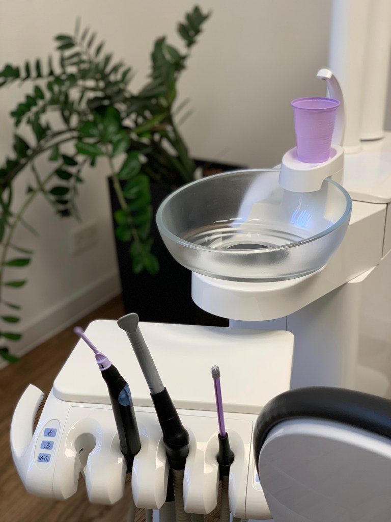 Behandlunsgzimmer bei der Zahnärztin: Blick auf den Wasserbecher, das Spuckbecken, die Geräte und ein Stückchen vom Zahnarztsessel. Im Hintergrund eine Palme. Alles, wie man es kennt, bis auf zwei Besonderheiten: Der Plastikbecher und die Dinger, aus denen die Luft zum Pusten kommt, sind lavendelfarbenen. Und zweitens ist das Spuckbecken aus nahezu durchsichtigem Glas. 