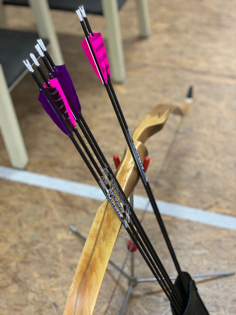 Sechs meiner Pfeile mit lila und pinken Federn in einer Köchertasche aus schwarzem Leder, die hinten in die Hosentasche passt. Die Pfeile lehnen an meinem Bogen, der wiederum mit der Sehne nach unten auf einem Bogenständer ruht. Das Ganze steht hinter einer weißen Linie auf dem Boden. 