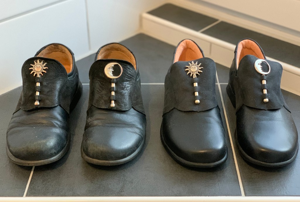 Das alte, abgetragene und das neue, unbenutzte Paar Schuhe. Es sind schwarze Glattlederschuhe zum Reinschlüpfen. Auf dem Spann haben sie eine Wildlederpasse, in der ein Silberstift steckt. Oben an diesem Stift, also Richtung Bein, ist am rechten Schuh eine silberne Sonne und am linken ein silberner Halbmond (durch einen Silberrand geschlossen zu einer kreisrunden Spange), beide mit Gesicht.