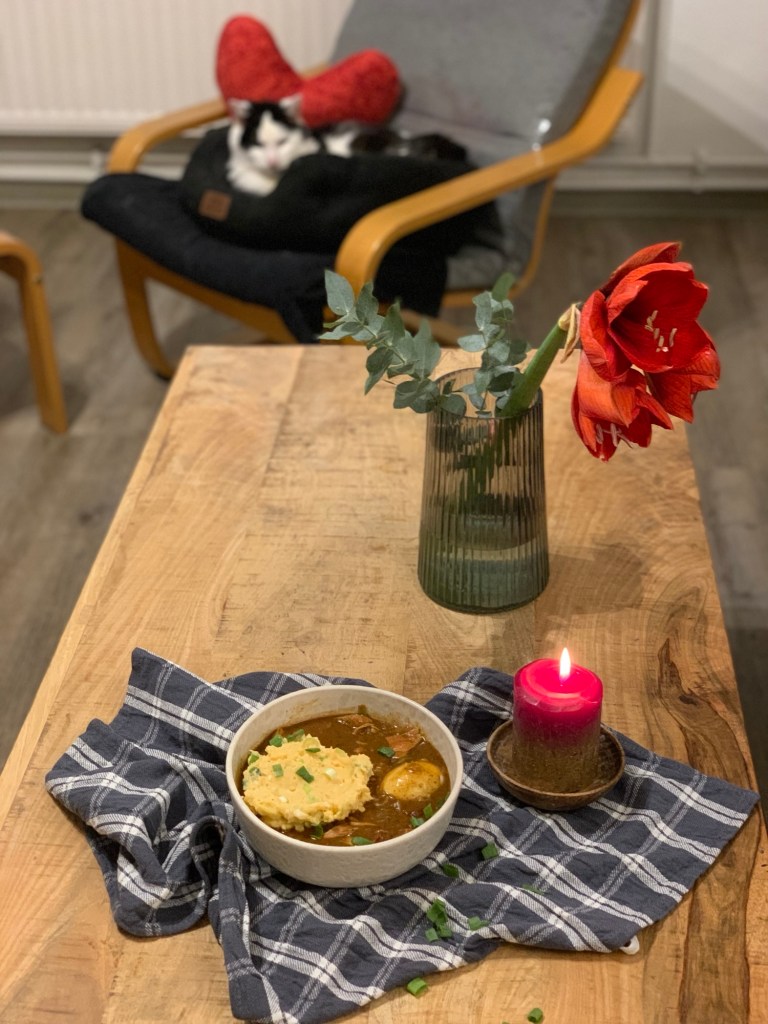 Vorne auf der Längsseite des Wohnzimmertisches auf einem graukarierten Küchenhandtuch eine Schale Gumbo mit Kartoffelsalat und einem Ei. Daneben eine brennende rote Stumpenkerze. Dahinter in einer Vase eine rote Amarillys mit drei Blüten. Im Hintergrund auf dem Pöang in seinem Katzenbettchen neben einem roten Stoffherz der #DiätKater.