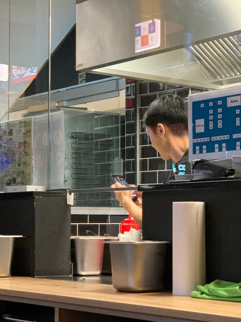 Der Kellner in Rückenansicht beim Filmen mit dem Handy. Die Küche ist durch Glasscheiben einsehbar. Unter den Trennscheiben ist an einer Stelle eine Lücke als Durchreiche.