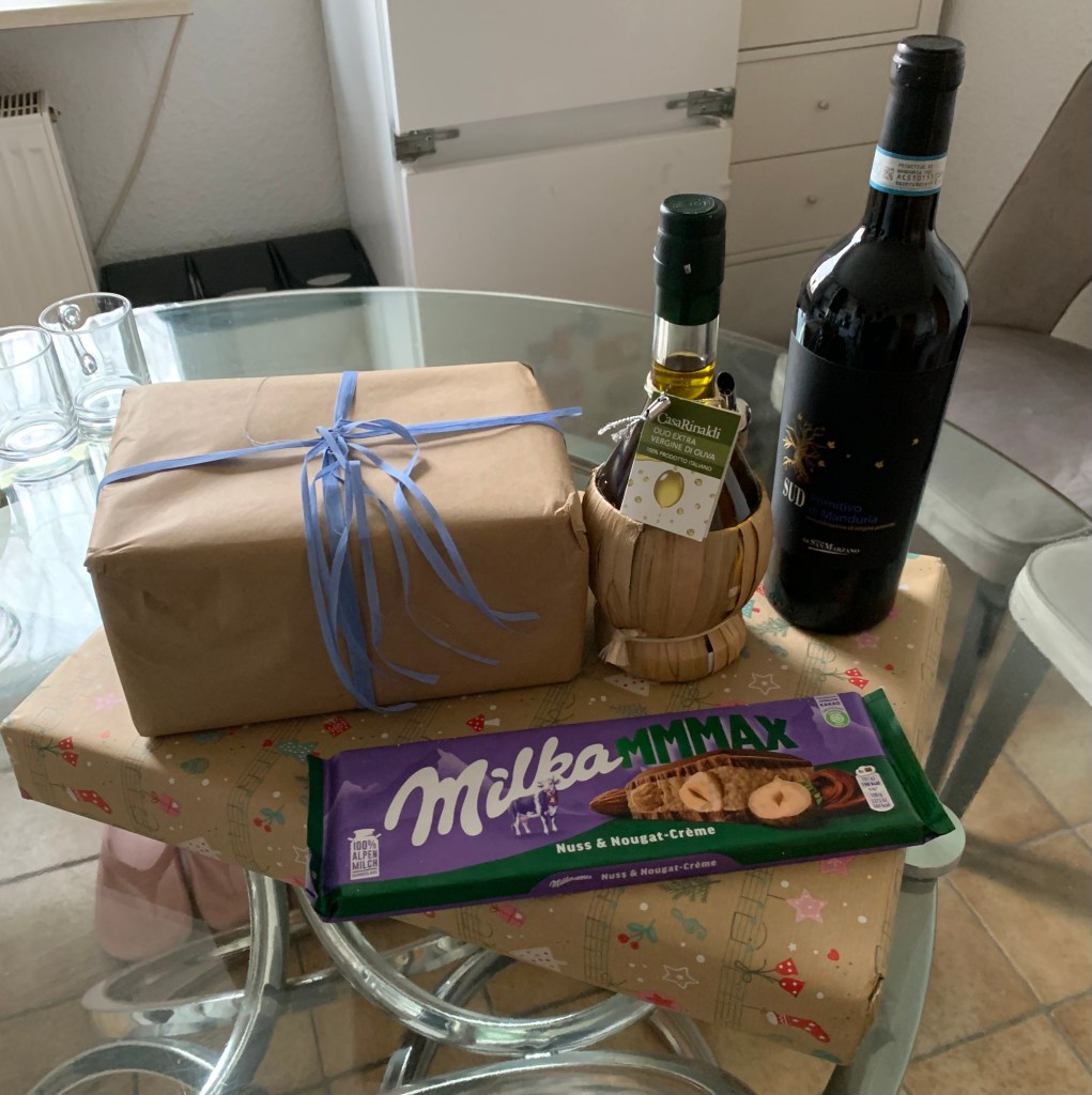Auf dem Glasküchentisch von Frau K. stehen meine Geschenke: Zwei Päckchen, eine Milka Max "Nuss & Nougat-Crême", eine Korbflasche gutes Olivenöl und eine Flasche Rotwein.