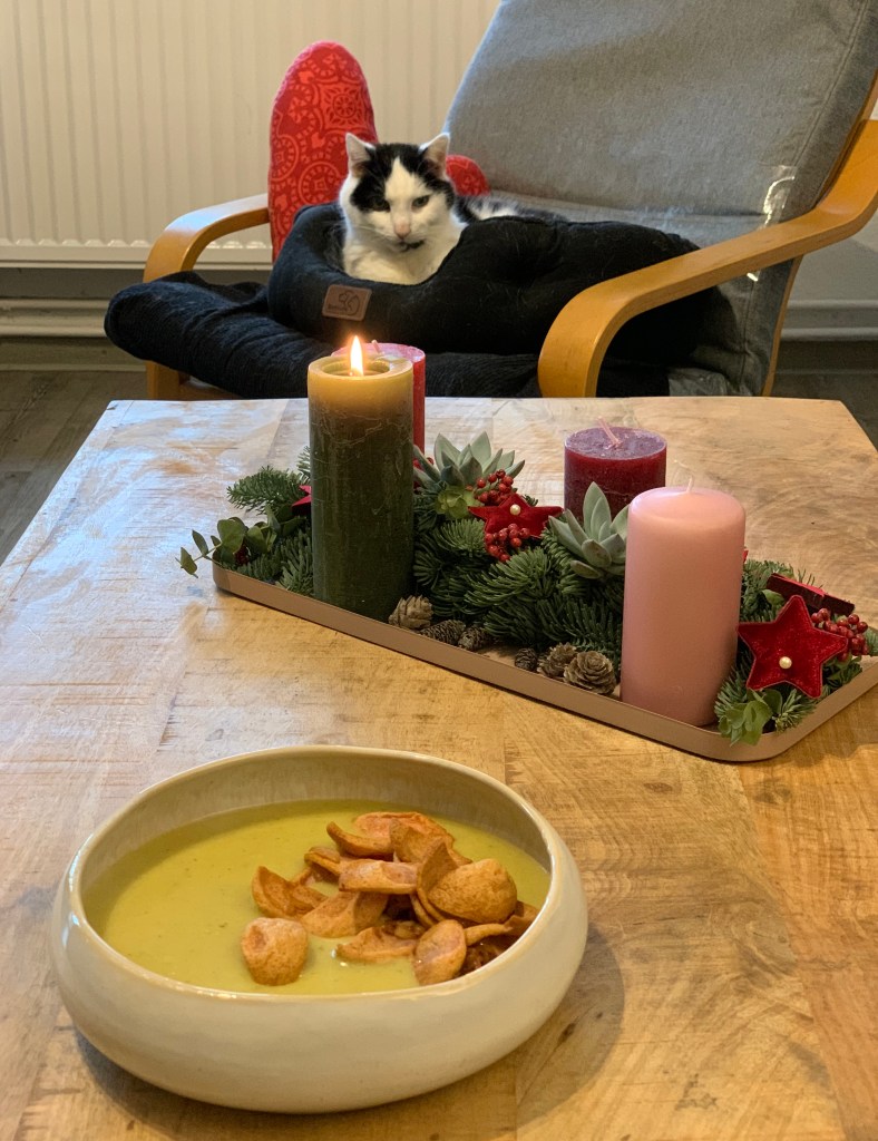 Vorne im Bild eine Schale Kartoffelsuppe mit Fleischwurst und Zwiebeln (sieht man nicht, da ganz wenige) als Topping. Dahinter mein Adventsgesteck mit brennender Kerze. Dahinter der #DiätKater in seiner Residenz, also seinem Katzenbettchen, auf dem Pöang (Ikeasessel).