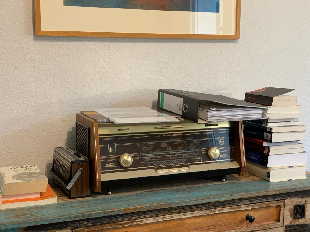 Blick auf ein historisches Radio in meinem Arbeitszimmer (Philips Bi-Ampli). Links daneben drei Bücher und ein kleines, historisches Kofferradio (Teddy 4, Schaub-Lorenz). Auf dem großen Radio ein paar Blätter in einer Plastikhülle und ein Leitzordner. Rechts daneben stapeln sich ein dutzend Bücher.