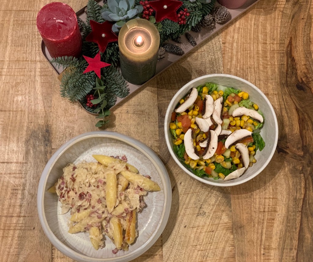 Mittagessen wie beschrieben, Schupfnudeln und Salat jeweils in einer Schale. Außerdem sieht man einen Teil des Adventsgestecks mit brennender Kerze.