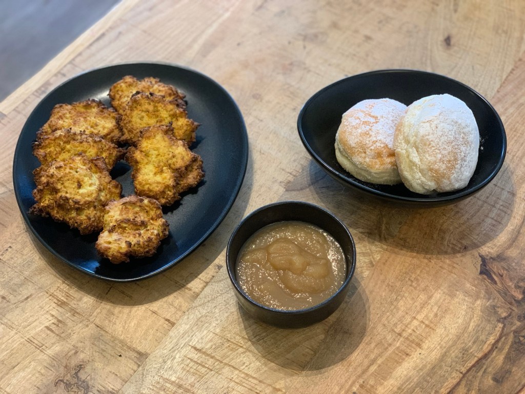 Sieben Reibeplätzchen auf einem oval-eliptsichen Teller, daneben eine ebenso geformte Schale mit zwei Berlinern und dazwischen ein rundes Schälchen mit Apfelmus. Alles drei aus derselben schwarzen Keramikserie.