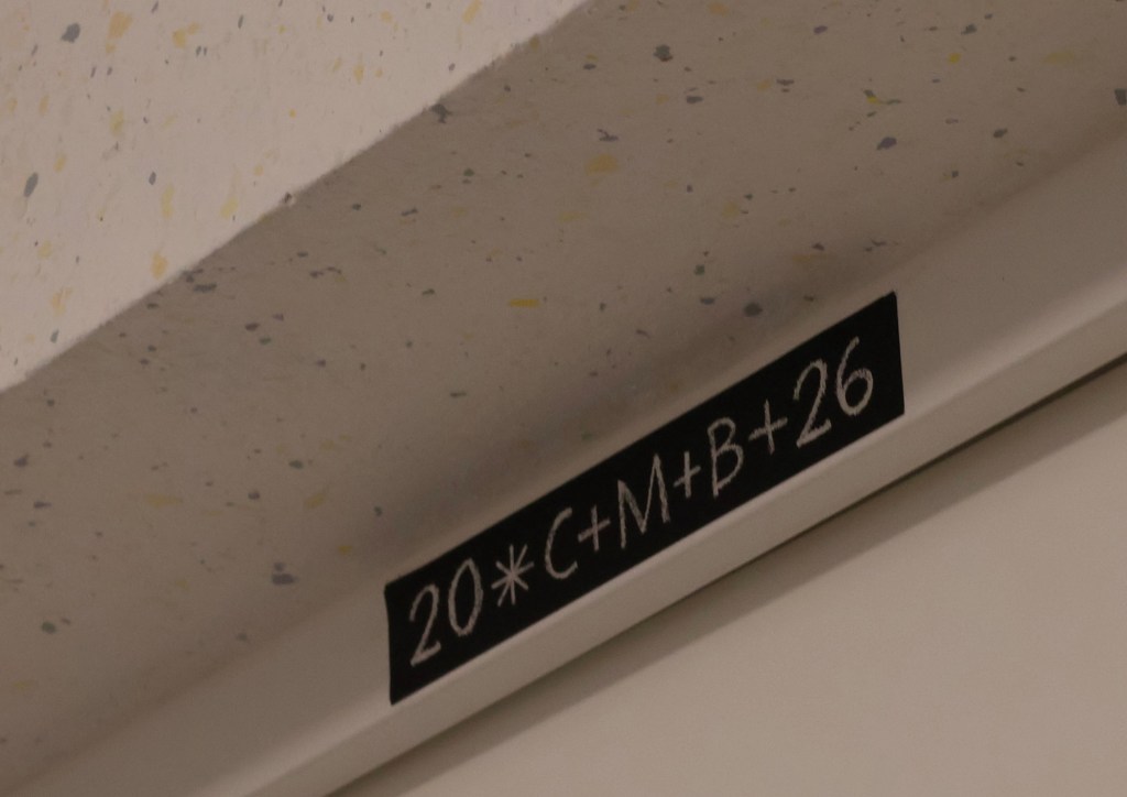 Klebesegen über der Wohnungstür. Man sieht von der Tür nur einen kleinen Ausschnitt, darüber den Segen und darüber der Sturz mit weißer Tapete mit dezenten gelben und grauen Punkten.