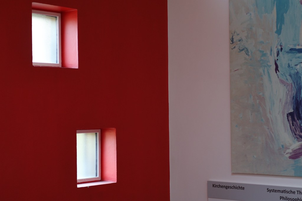 Blick auf den Winkel des Treppenhauses, an dem zwei Wände zusammentreffen. Von der Treppe sieht man dabei nichts. Die linke Wand ist kpmplett knallrot gestrichen und verfügt über zwei quadratische Fenster mit Milchglasscheiben. An der rechten Wand hängt ein modernes, abstraktes Kunstwerk in hellen Blautönen, von dem man hier einen Ausschnitt sieht. Darunter, ebenfalls als Auschnitt, ein Schild zur Orientierung. Das Wort "Kirchengeschichte "ist ganz, die Wörter "Systematische Theologie" und "Philosophie" sind in Teilen zu lesen.