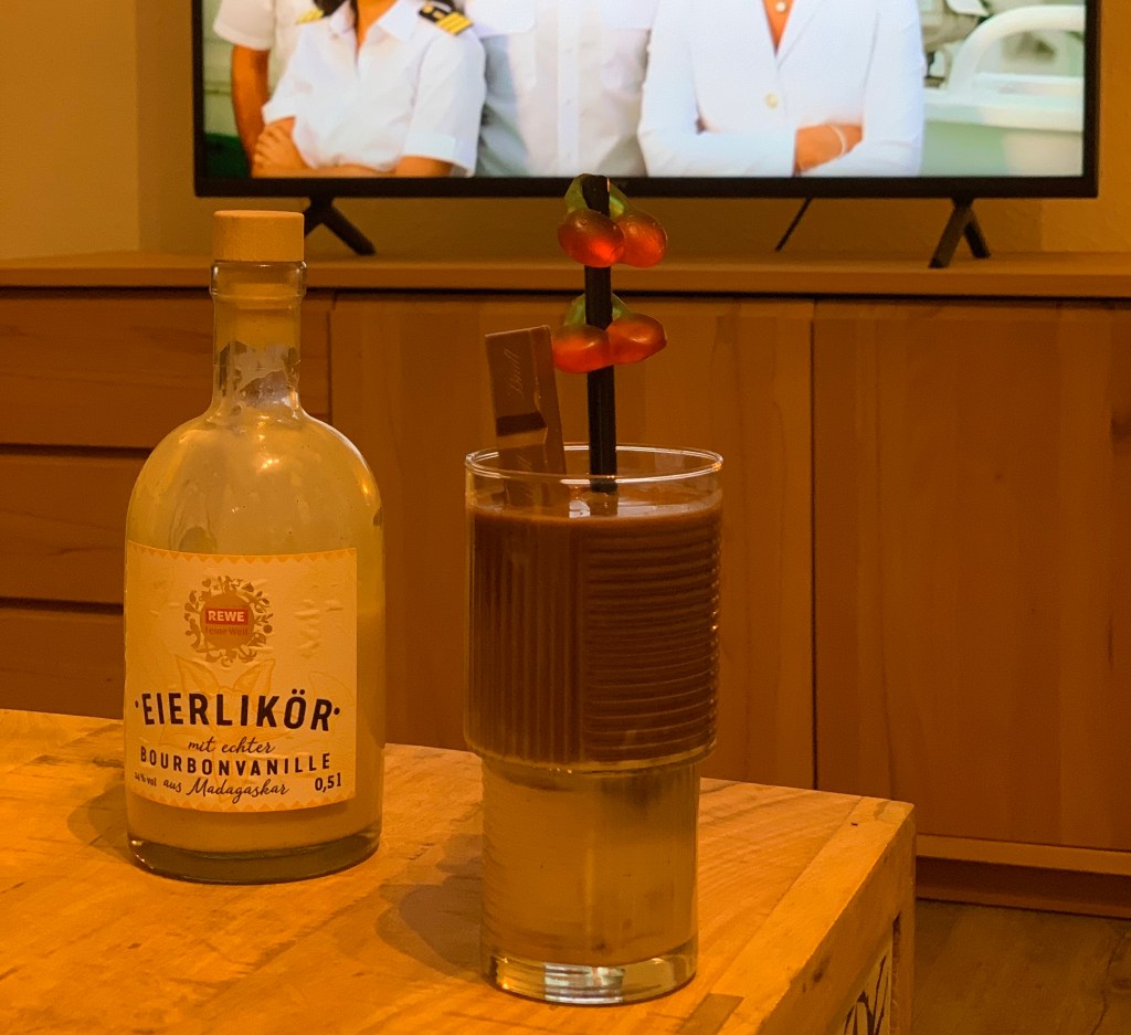 Eine Flasche Eierlikör von Rewe, daneben ein Glas mit unten Eierlikör, oben Kakao, einem Strohhalm aus Metall mit Haribokirschen und einem Lindt-Schokoriegel darin. Im Hintergrund das TV, wo gerade die vier Hauptdarsteller:innen vom Traumschiff eingeblendet sind. Aber ich habe ihnen aus urheberrechtlichen Gründen die Köpfe abgeschnitten, also den Bildrand oben verkürzt.