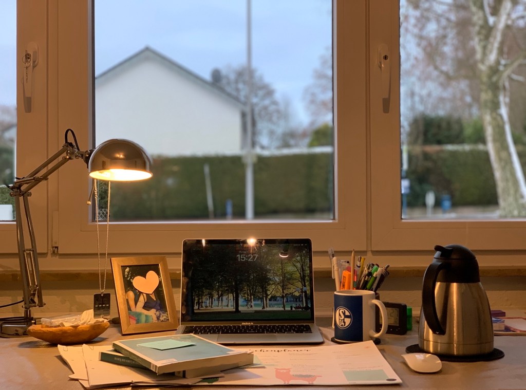 Blick auf meinen Schreibtisch mit Lampe, Büchern, Texten, Macbook, Tee (Schalke-Tasse und Kanne), Stiften usw. Dahinter die Fenster mit Blick Richtung Straße, Nachbarhecke und -haus.