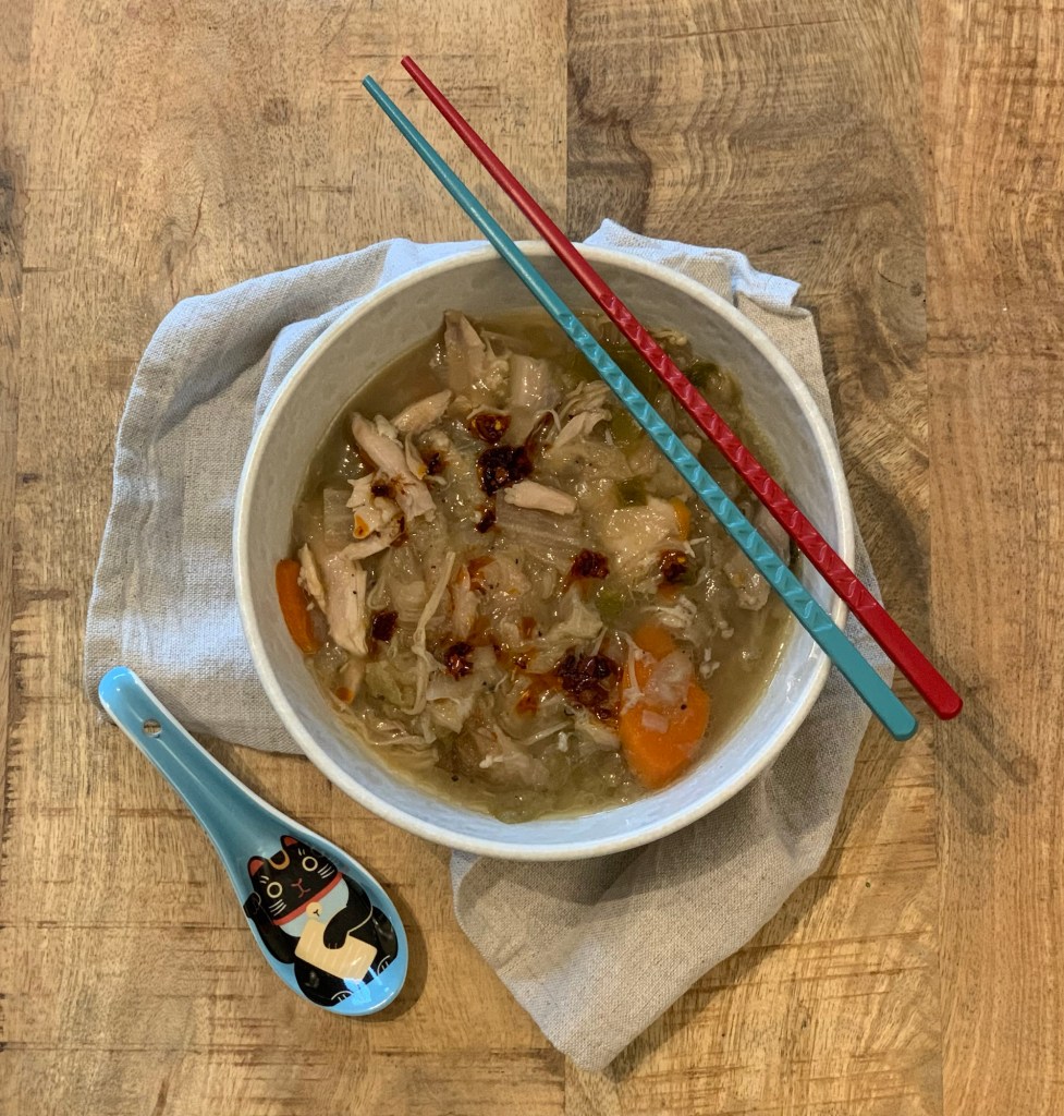 Eine Portion Suppe in einer cremefarbenen Schale auf einer Leinenserviette. Auf der Schale liegen ein blautürkises und ein rotes Ess-Stäbchen. Neben der Schale liegt ein chinesischer Suppenlöffel, ebenfalls in türkisblau, in dessen Laffe eine schwarze Winkekatze mit roten Ohren aufgedruckt ist.