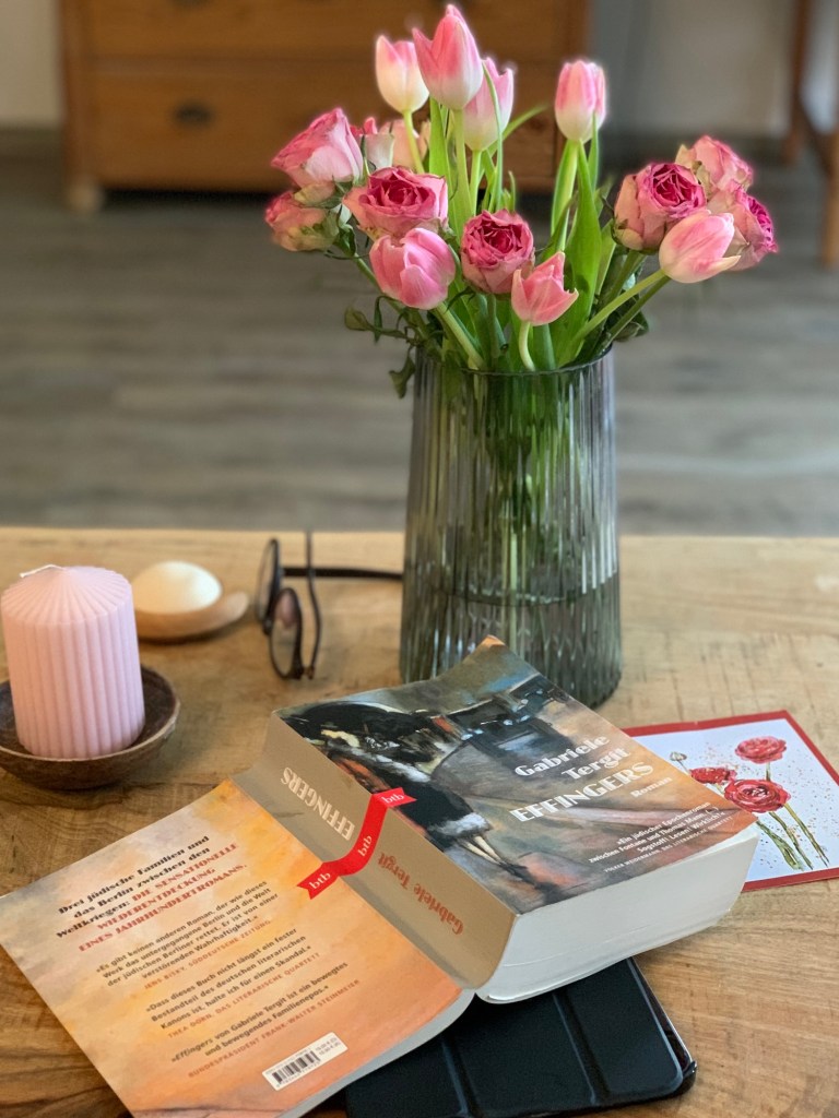 Blick auf das gewollte Chaos auf meinem Wohnzimmertisch: Ein Strauß rosa Tulpen und Rosen in einer braungeriffelten Glasvase, links daneben eine noch nicht angezündete rosa Stumpenkerte, ein Duftstein und meine Brille. Davor das aufgeklappte Taschenbuch mit Titel und Rüclen nach oben. Rechts daneben eine Postkarte von Wandklex mit roten Rosen, die ich als Lesezeichen benutzt habe. Unter dem Buch guckt man iPad mini hervor, in einer zugeklappten, schwarzen Hülle.