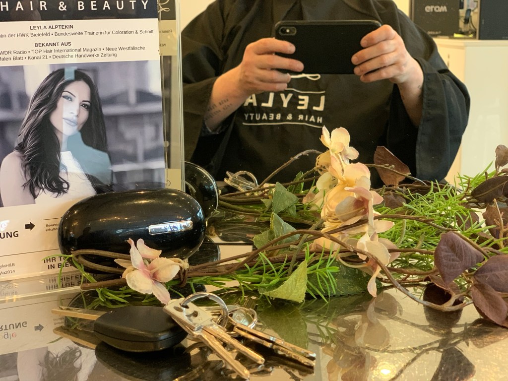 Bei der Friseurin vor mir auf der Ablage ein kleiner Werbe-Aufsteller mit dem Bild einer langhaarigen Frau, meine Autoschlüssel, mein Oberkörper mit schwarzem Umhang, davor halte ich das Handy.