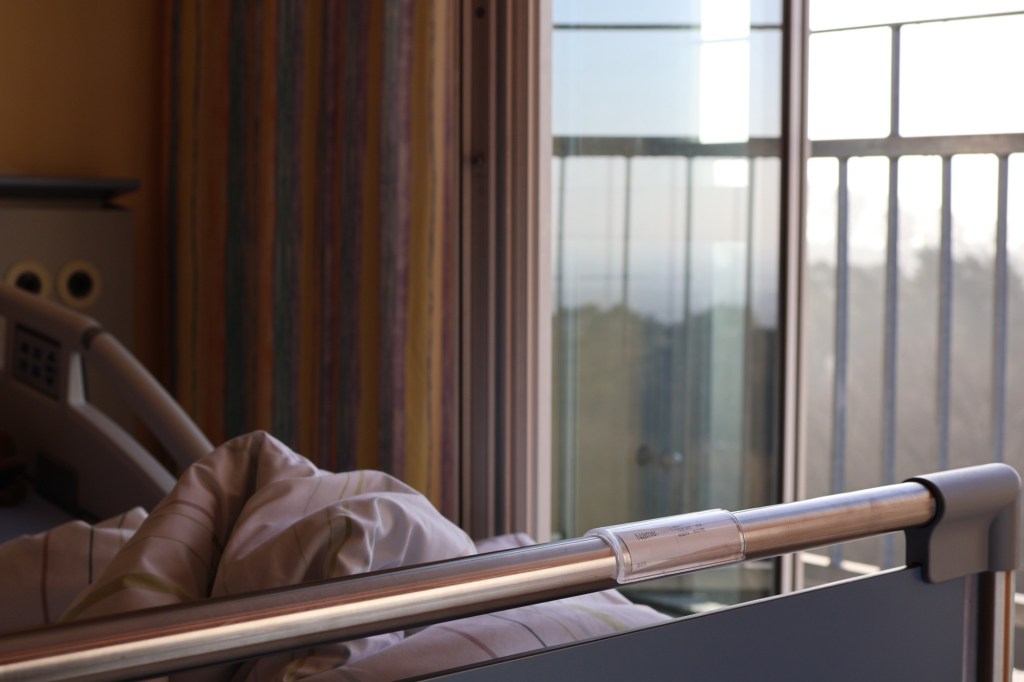 Mein Krankenbett mit zerwühlter Bettdecke, im Hintergrund das geöffnete Fenster mit Sicherheitsbalkongitter und dem schemenhaften Blick auf den Teutoburger Wald. (Aus meiner Reihe: Geöffnetes Fenster am Morgen.)