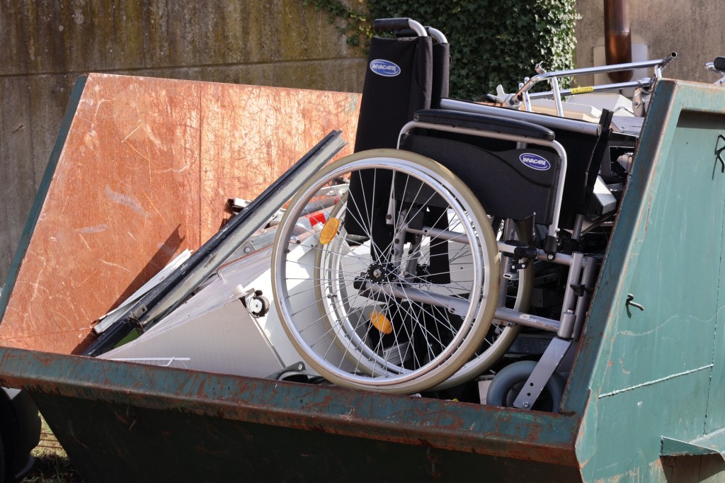 Ein Schrottcontainer mit Metallabfällen eines Krankenhauses. Zuvörderst sichtbar ein Rollstuhl.