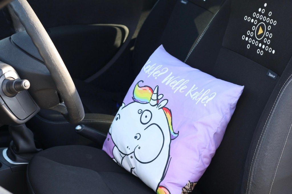 Blick auf den Fahrersitz meines Autos. Dort liegt ein fliederfarbenes, fluffiges Kissen von der Größe eines Sofakissens, auf dem ein moppeliges, kaugendes Einhörn abgebildet ist. Darüber steht in weißer Schrift "Kekfe? Welfe Kekfe?"