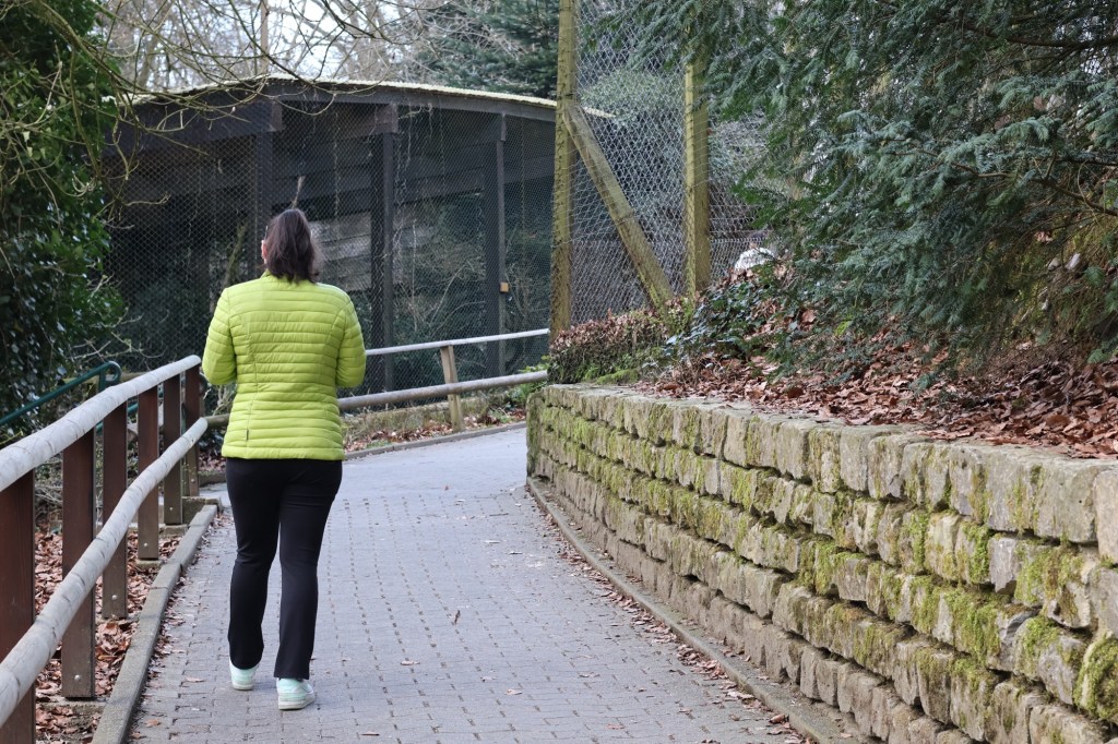 Frau K., in grüner Jacke und schwarzer Hose, geht den Fußweg in Sichtweise zweier Gehege entlang (Rückenansicht).