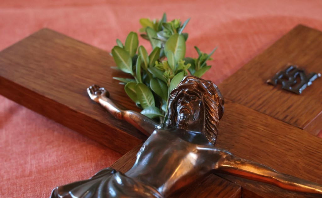 Das Kruzifix liegend auf einer dunkelorangenen Tischdecke. Hinter der rechten Schulter Jesu steckt ein kleiner, frischer Buchsbaumzweig.