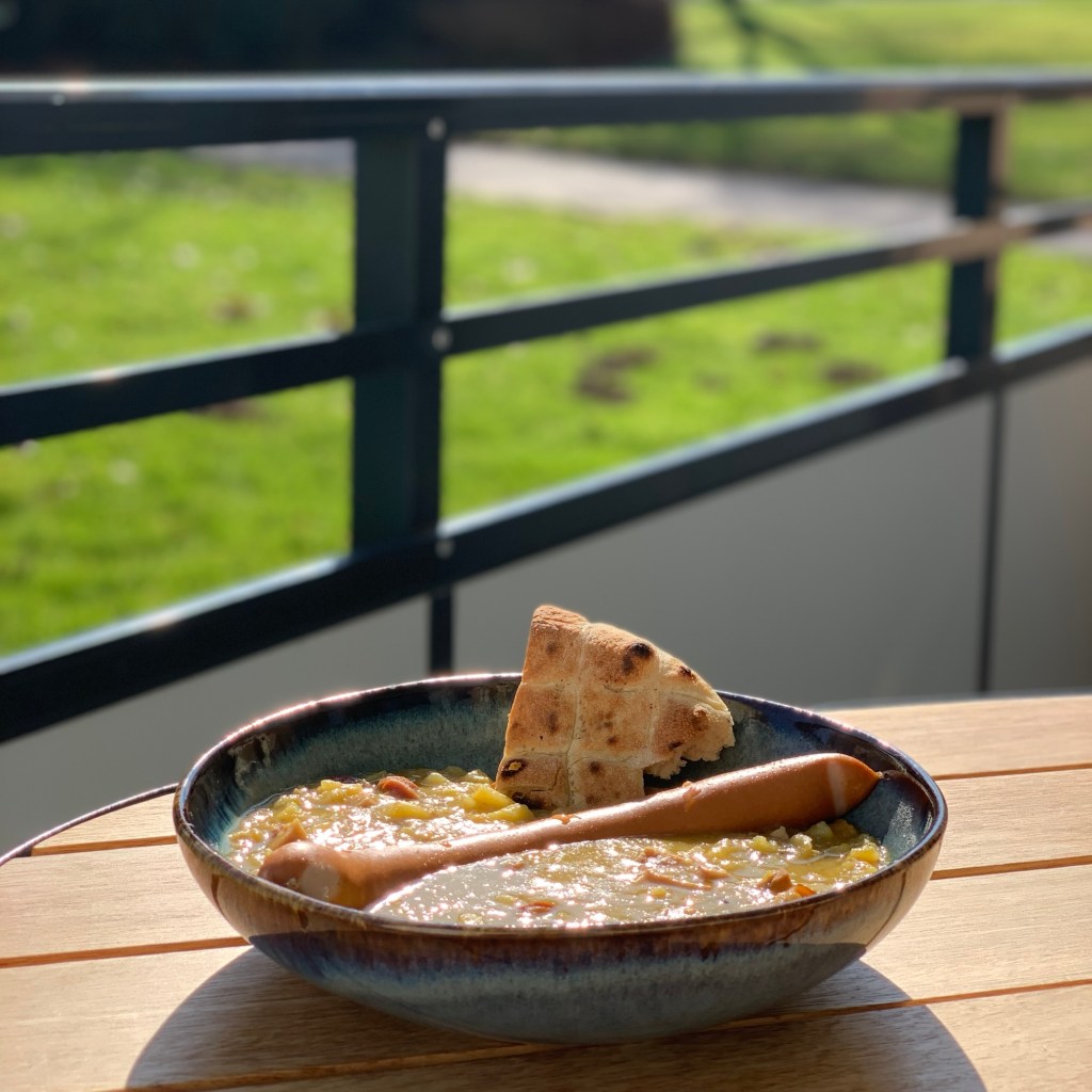Der Teller mit Suppe, Würstchen und Brotstück auf dem Balkontisch. Im Hintergrund verschwommen der Blick auf Gebüsch, Wiese und Straße. Man erahnt einige Maulwurfshügel.