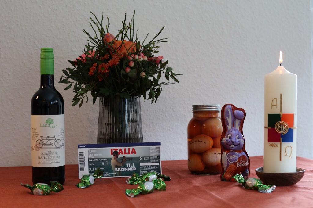Mein Ostergabentisch mit Blumenstrauß und brennender Osterkerze. Dazu außerdem: Eine Flasche Dornfelder Rotwein (trocken), eine Eintrittskarte zu Till Brönner, ein Glas Soleier, ein Milka-Lotus-Osterhase, fünf Schokokrokanteier von Lindt.