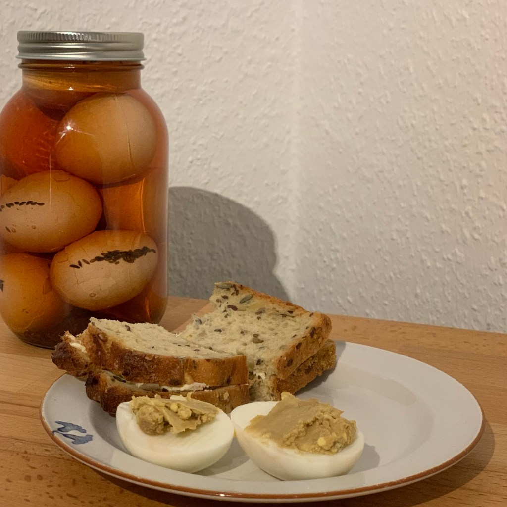 Ein Schraubglas mit Soleiern und davor ein Teller, darauf eins zubereitet und ein geklapptes und halbiertes Butterbrot.