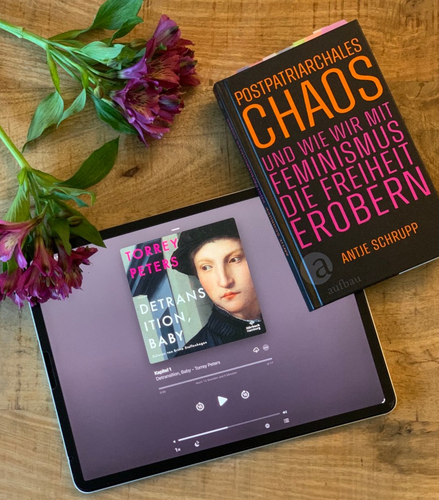 Das Hörbuch auf meinem iPad pro und das Papierbuch, zusammen mit zwei lila Dolden Fresien, passend zur Farbgebung der Bücher. Aus dem Papierbuch gucken oben die ganzen Einmerker raus.