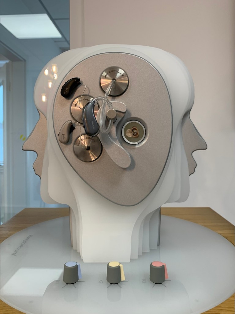 Ein stilisierter Januskopf aus Edelstahl und weißem Kunststoff mit Technikkrams und drei Hörgeräten sowie drei Drehknöpfen auf der Basis vor dem Hals.